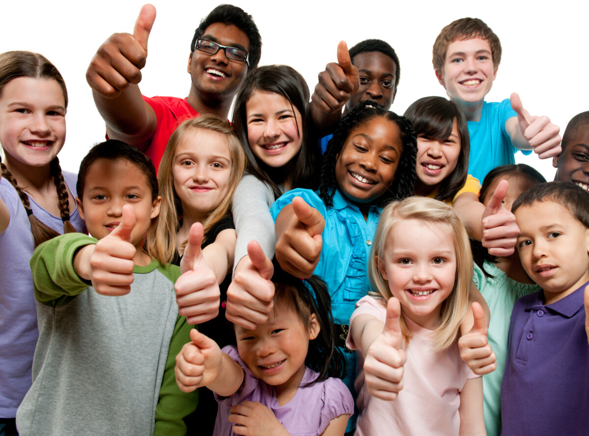 A group of K through grade 12 children standing together giving a thumbs up. The children are all different ages and races. The children have on purple, blue, red and gray shirts, are all smiling and standing in front of a white background.