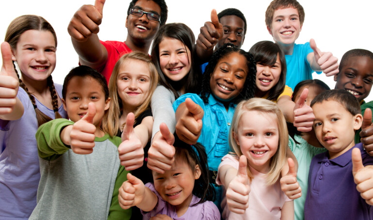A group of K through grade 12 children standing together giving a thumbs up. The children are all different ages and races. The children have on purple, blue, red and gray shirts, are all smiling and standing in front of a white background.